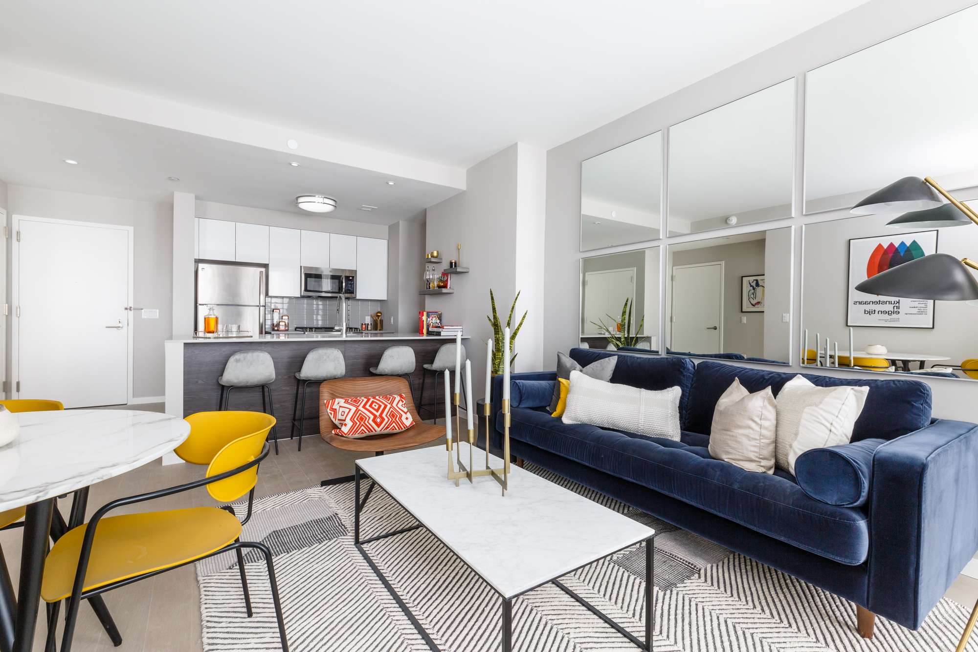 Open layout with living room with blue couch and white coffee table, round dining table, mirrors on walls and galley kitchen with stainless steel appliances and bar stool seating counter near entrance