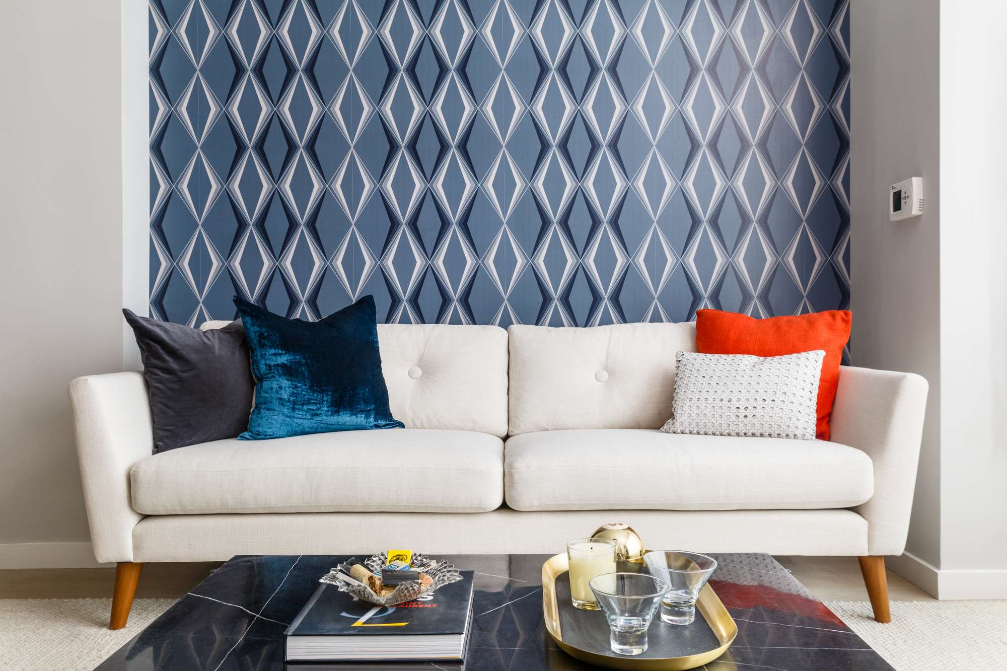 Living room detail with white couch, decorative pillows, blue pattern accent wallpapered wall and coffee table with book, candle and drinks
